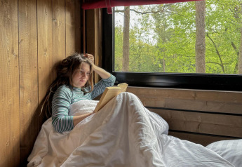 Donna che legge un libro a letto accanto a una grande finestra con vista sulla foresta, Comfort Classic, Belgio.