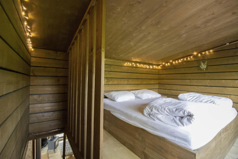 Cama tipo loft con ropa de cama blanca, luces y paredes de madera en una cabaña con jacuzzi en Limburg, Bélgica.
