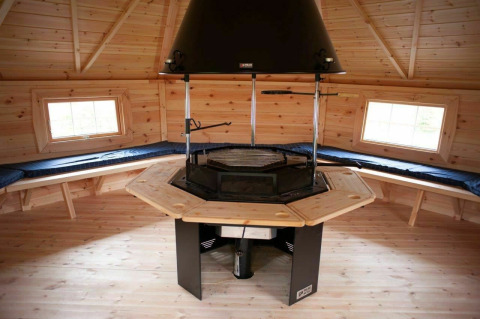 Interieur van een houten lodge met centrale barbecue en banken in Veluwe Villa + sauna, De Boshoek, Nederland.