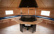 Interior de cabaña de madera con parrilla central y bancos en Veluwe Villa + sauna, Holiday park De Boshoek, Países Bajos.