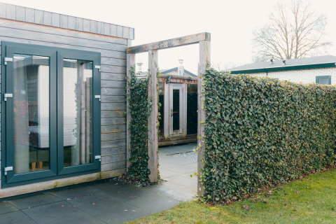 Buitenruimte van Veluwe Villa + sauna in vakantiepark De Boshoek, met haag en houten gebouwen.