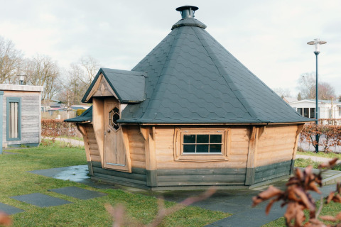 Kleine houten lodge met zwart dak aan vakantiepark De Boshoek in Nederland, omgeven door gras.