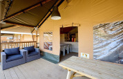 Outdoor sitting area of a luxury safari tent featuring cushioned wicker armchairs and a wooden picnic table.