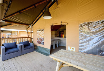 Espace salon extérieur d'une tente safari de luxe avec fauteuils confortables et table en bois sur la terrasse.