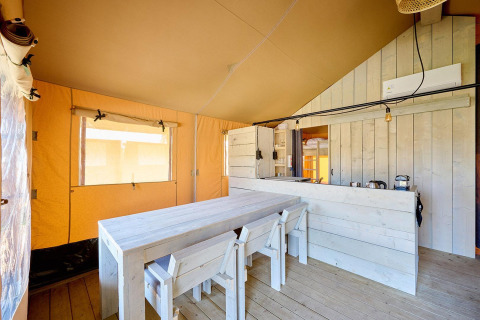 Vista interior de una tienda safari con mesa de madera, sillas y una pequeña cocina al fondo.