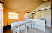 Interno di una tenda safari con tavolo di legno, sedie e un piccolo angolo cottura sullo sfondo.