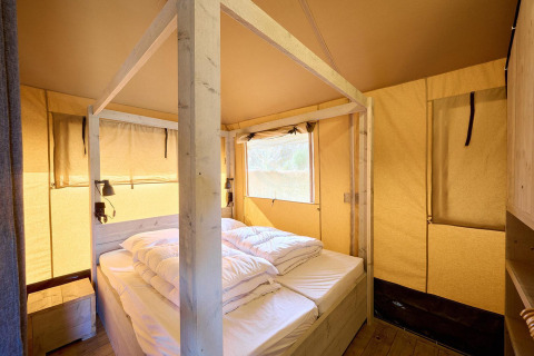 Interior view of a safari tent featuring a cozy double bed, wooden frame, and warm natural lighting.