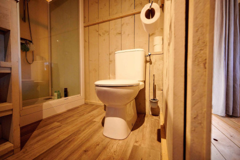 Baño en tienda safari con inodoro moderno, paredes de madera, ducha y suelo de madera natural.