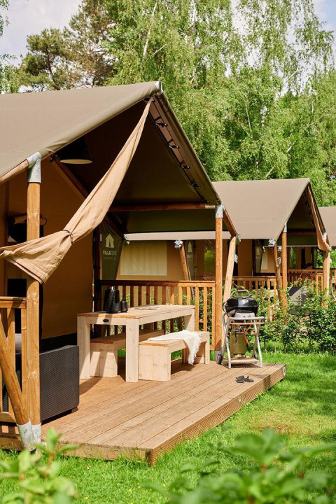 Tienda safari con terraza de madera, mesa de picnic y barbacoa, rodeada de vegetación y árboles verdes.