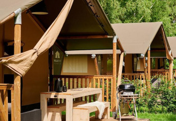 Tenda safari con veranda in legno, tavolo da picnic e barbecue, immersa nel verde e tra gli alberi al sole.