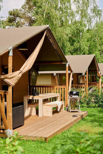 Tienda safari con terraza de madera, mesa de picnic y barbacoa, rodeada de vegetación y árboles verdes.
