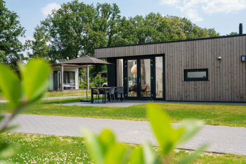 Moderna cabaña de madera con terraza, muebles de jardín y sombrilla, rodeada de césped y árboles.
