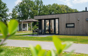 Moderne lodge med træfacade, terrasse, havemøbler og parasol, omgivet af grønt græs og træer.