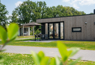 Lodge moderna in legno con terrazza, tavolo da esterno e ombrellone, circondata dal verde e alberi.