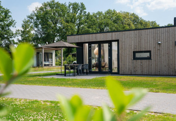 Moderne lodge med træfacade, terrasse, havemøbler og parasol, omgivet af grønt græs og træer.