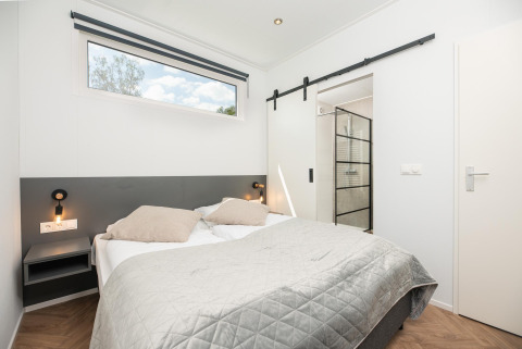 Modern lodge slaapkamer met tweepersoonsbed, wandlampen, nachtkastjes en glazen deur naar badkamer.