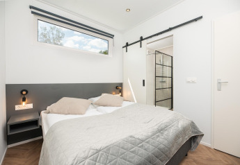 Slaapkamer in een moderne lodge met dubbel bed, nachtlampjes en glazen schuifdeur naar de badkamer.