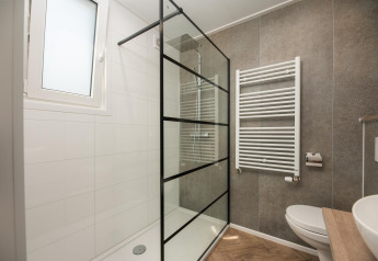 Moderne lodgebadeværelse med glasbrusevæg, hvid radiator, toilet og trævask i minimalistisk stil.