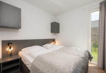 Chambre de lodge moderne avec lit double, armoires grises, lampes de chevet et grande fenêtre sur jardin.