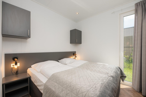 Moderne lodge slaapkamer met dubbel bed, grijze kasten, nachtlampjes en een groot raam naar de tuin.