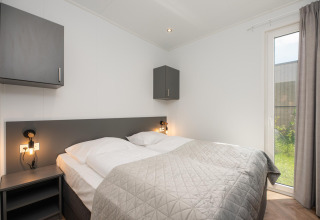 Moderne lodge slaapkamer met dubbel bed, grijze kasten, nachtlampjes en een groot raam naar de tuin.