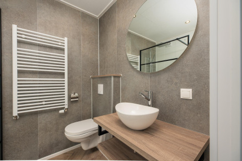 Moderne lodgebadkamer met hangtoilet, houten blad, ronde waskom, grote ronde spiegel en grijze tegels.