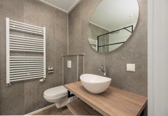 Salle de bain moderne d’un lodge avec WC suspendu, vasque ronde, miroir circulaire et carrelage gris.