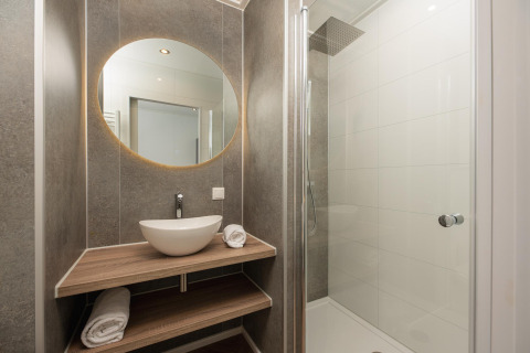 Baño moderno en una cabaña con espejo redondo, lavabo, estantes de madera y ducha con mampara de cristal.