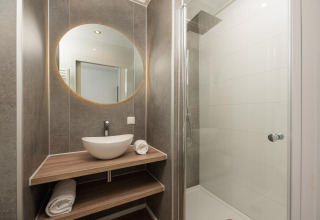 Baño moderno en una cabaña con espejo redondo, lavabo, estantes de madera y ducha con mampara de cristal.