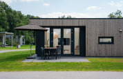Moderne lodge med træbeklædning, udendørs terrasse, parasol og spisebord på en grøn græsplæne.