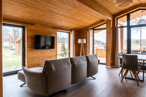 Salon moderne de lodge en bois avec canapé gris, grandes fenêtres et salle à manger au Lakehouse Scandi, Allemagne.