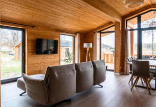 Moderne lodge-stue med trægulve og -vægge, store vinduer, grå sofa og spisebord i Lakehouse Scandi, Tyskland.