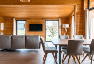 Moderne stue i Lakehouse Scandi, Wilsumer Berge, Tyskland, med træpaneler, sofa, spisebord og store vinduer.