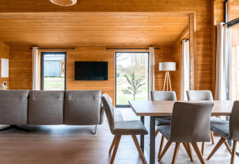 Moderne stue i Lakehouse Scandi, Wilsumer Berge, Tyskland, med træpaneler, sofa, spisebord og store vinduer.
