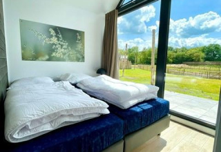Dormitorio luminoso con grandes ventanales en una tiny house en Camping Si-Es-An, Países Bajos, vista a la naturaleza.