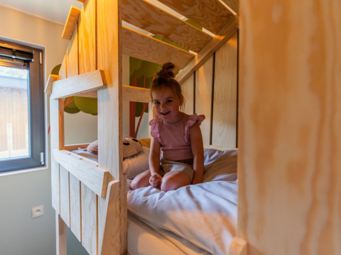 Kleines Mädchen sitzt lächelnd im Etagenbett der Kids cottage Lodge in Kaatsheuvel, Niederlande.