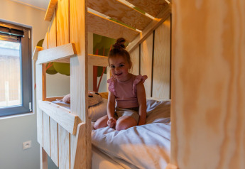 Lille pige sidder glad i en køjeseng på Kids cottage lodge i Kaatsheuvel, Holland med træindretning.
