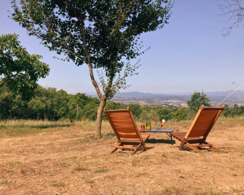Twee ligstoelen en een tafel onder een boom met uitzicht op de heuvels bij Camping Lucherino in Toscane.