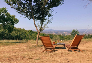 Two deck chairs and a table under a tree overlooking the hills at Camping Lucherino in Tuscany, Italy.