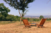 Two deck chairs and a table under a tree overlooking the hills at Camping Lucherino in Tuscany, Italy.