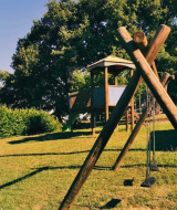 Playground at Camping Lucherino in Tuscany, Italy, featuring swings and a wooden playhouse on a hill.