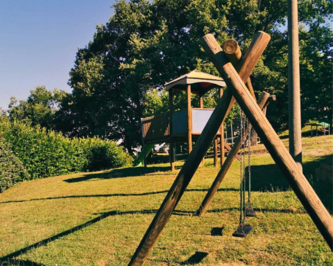 Parque infantil en Camping Lucherino, Toscana, Italia, con columpios y casita de madera en una colina verde.