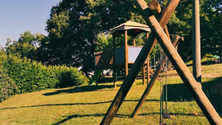 Parque infantil en Camping Lucherino, Toscana, Italia, con columpios y casita de madera en una colina verde.