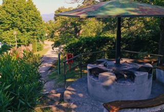 Buitenbarbecue met banken bij Camping Lucherino in Toscane, Italië, omgeven door bomen en wandelpad.