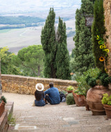 Dos personas se sientan en escaleras de Cinigiano, Toscana, rodeadas de macetas y un paisaje panorámico.