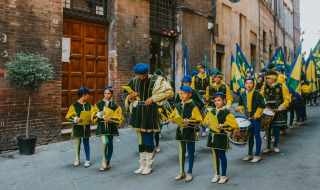 Desfile tradicional con niños y adultos en coloridos trajes y banderas en una calle de Cinigiano, Toscana, Italia.
