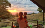 Una donna tiene in braccio un bambino e guarda il tramonto dalla Safari Tent Valley al Camping Lucherino, Italia.