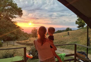 Una donna tiene in braccio un bambino e guarda il tramonto dalla Safari Tent Valley al Camping Lucherino, Italia.