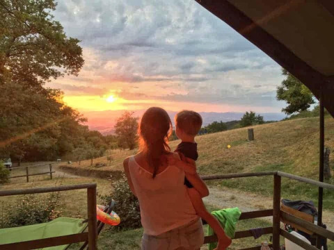 Una donna tiene in braccio un bambino e guarda il tramonto dalla Safari Tent Valley al Camping Lucherino, Italia.