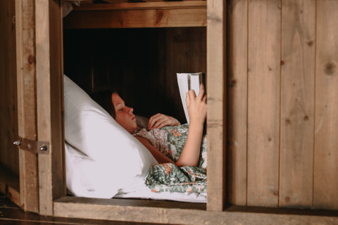 Ein Kind liegt in einer gemütlichen Holzkoje im Feather Down Rochefort-Ardenne in Belgien und liest ein Buch.